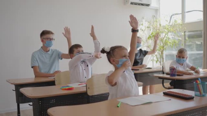 学校教室里戴着面具的孩子坐在教室里，以慢动作举手回答老师的问题。在学校大流行期间的课程