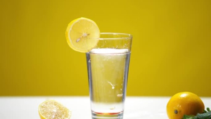 特写玻璃杯，里面有清爽的柠檬水，水滴和水果坑以慢动作落下。黄色背景下的有机维生素鸡尾酒制剂。