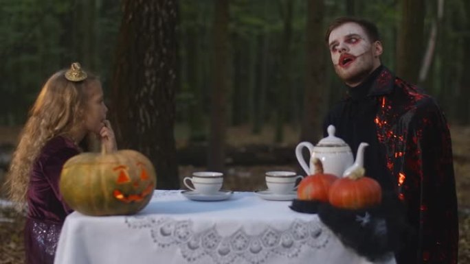 体贴的小女孩和穿着吸血鬼服装的男人坐在森林里的桌子上互相检查。万圣节在户外林地里的高加索孩子和实体。