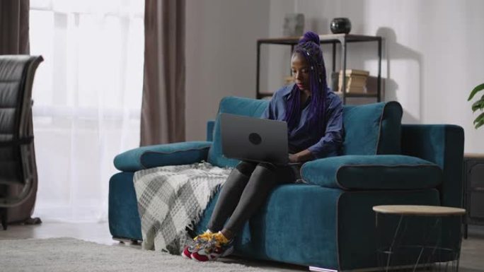 单身非洲裔美国妇女正在使用笔记本电脑，周末独自一人坐在家里，坐在房间的沙发上上网
