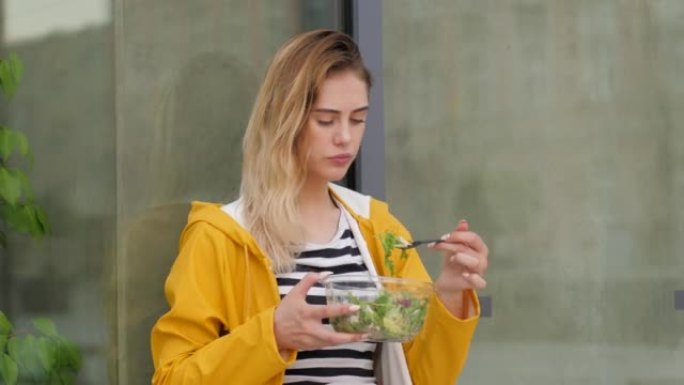 美丽的年轻女孩吃塑料碗里的健康蔬菜沙拉，穿着黄色雨衣独自站在城市里，靠在玻璃墙上