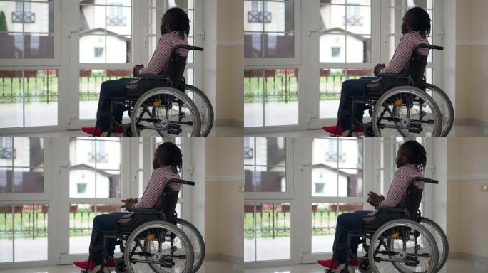 坐在轮椅上的宽镜头体贴的残疾非裔美国人坐在室内看着窗外。客厅里年轻英俊瘫痪的人在家思考的肖像。