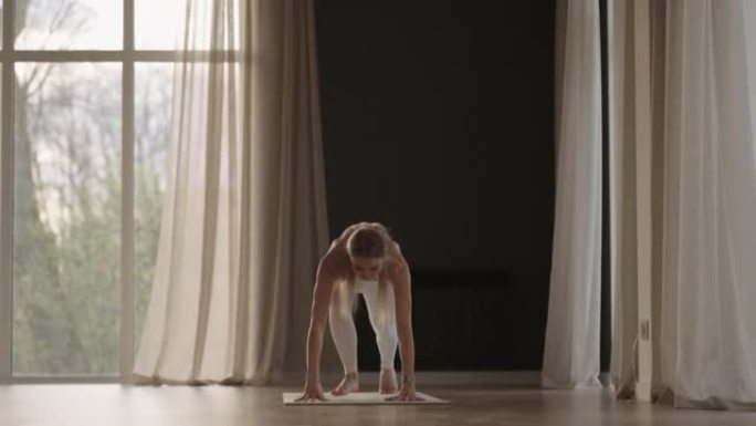 一名穿着白色西装的年轻女子在健身房以慢动作在阳光下训练瑜伽