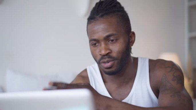 积极的非洲裔美国男性自由职业者使用笔记本电脑上的虚拟会议交谈和微笑。英俊的年轻人在室内家庭办公室里在