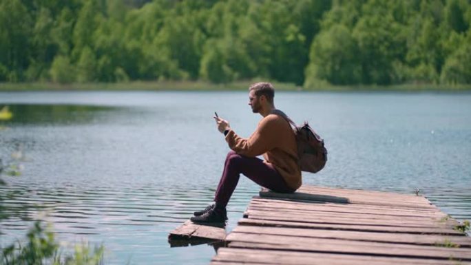 一位戴着眼镜、背着背包、蓄着胡须的年轻男子坐在湖边的一个木制码头上，用慢镜头打印出一条信息，用手机铺