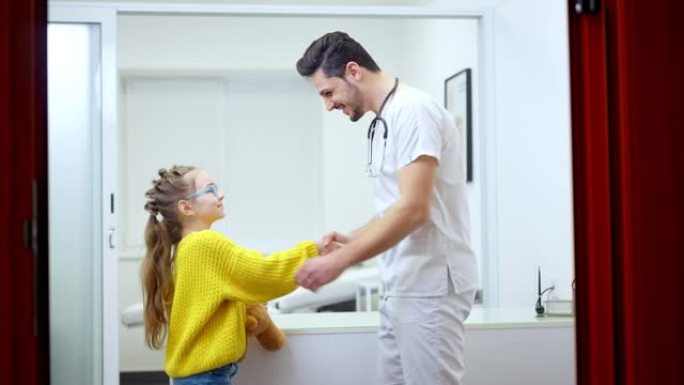 快乐的医生和小病人在诊所握手的侧视图。开朗的中东男人和可爱的高加索女孩站在医院里说话和微笑。儿科和医