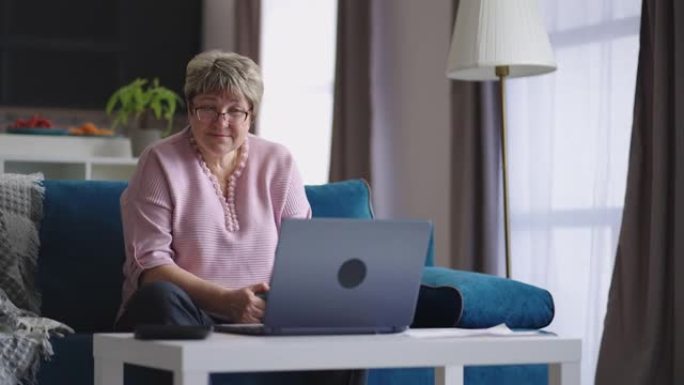 老年妇女正在通过视频通话使用笔记本电脑进行在线交流，女性退休人员坐在家里的沙发上
