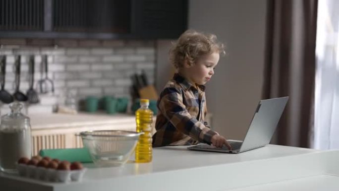 周日早上，迷人的卷发小男孩在厨房用笔记本电脑观看视频，中等肖像