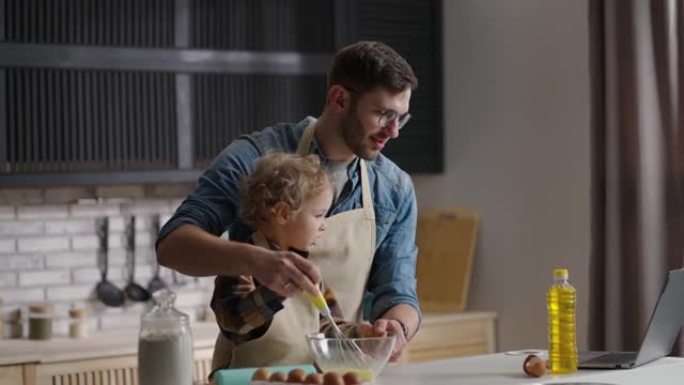 小儿子和父亲正在观看笔记本电脑上的教程，在家庭厨房做饭，在碗里打鸡蛋，爸爸和孩子