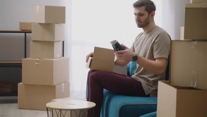 一个年轻人打开一个纸箱，搬走后在新公寓里拿出坐在沙发上的东西。购买新公寓