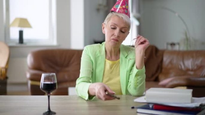 戴着派对帽的无聊悲伤的高加索中年妇女的肖像，检查电话留言，慢动作喝红酒。孤独优雅的女士在室内家庭办公