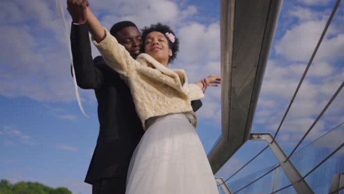快乐微笑的新婚夫妇举手模仿泰坦尼克号的场景，背景是夏天的天空。激动深情的非洲裔美国新娘和新郎享受婚姻
