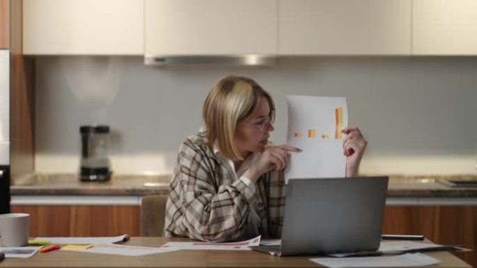 一位在家中戴眼镜的年轻女子坐在厨房里，对着笔记本电脑的相机展示了一张图表。家庭办公室远程工作。用图表