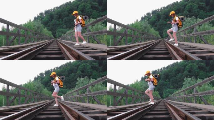 一位女游客背着背包沿着桥上的铁轨行走。一个带着橙色背包、帽子和相机徒步旅行的女孩。积极旅游生活方式的