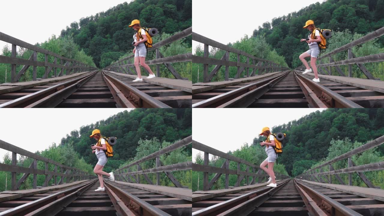 一位女游客背着背包沿着桥上的铁轨行走。一个带着橙色背包、帽子和相机徒步旅行的女孩。积极旅游生活方式的