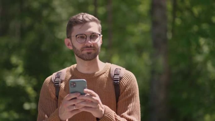 一个背着背包的男性旅行者沿着森林中的一条土路行走，写信息，在互联网上交流，查看内容，在手机上做笔记，