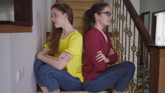 争辩说苗条美丽的同卵女人背靠背坐在楼梯上叹息。迷人自信的时尚女士和书呆子兄弟姐妹在室内。个性概念。
