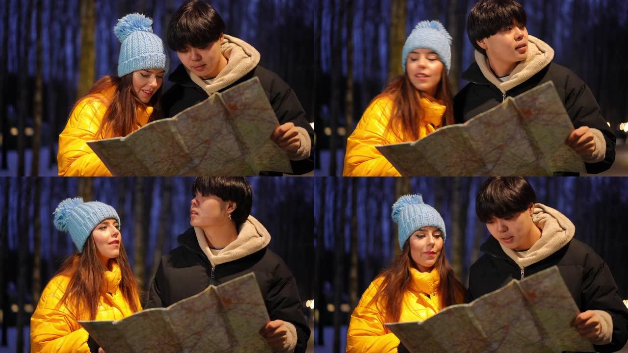 失落的亚洲男人和白人女人在冬天的夜晚站在户外，看着周围的纸地图。不确定几个年轻游客选择路线。导航和旅