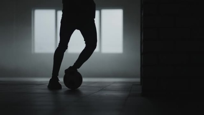 一名足球运动员在太阳和升起的灰尘下在地下停车场训练的剪影。体育抱负和毅力的成功概念。自由式足球