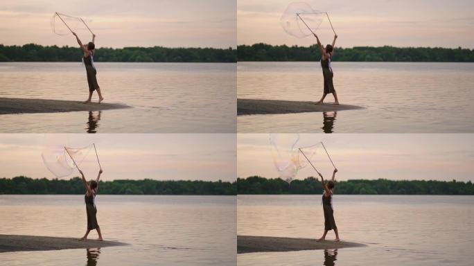 一个年轻的嬉皮士女人穿着裙子，头上长着羽毛，在日落时在湖岸慢动作时产生巨大的肥皂泡