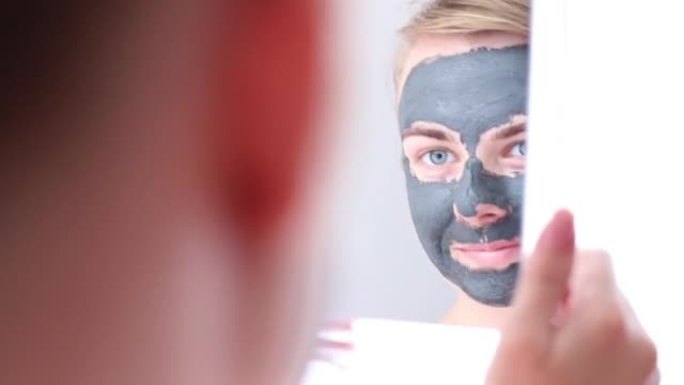 一个脸上戴着粘土制成的化妆品面膜的女人照镜子。家庭护肤。美容护理