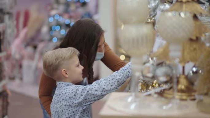 一位戴着医用口罩的母亲和她的儿子在购物中心为圣诞节选择家居装饰品