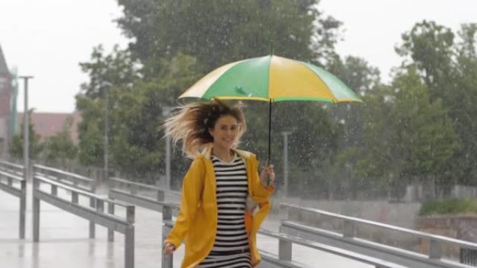 慢动作可爱的年轻女孩穿着黄色雨衣打着雨伞在淋浴雨中奔跑