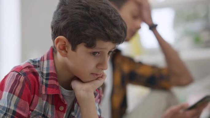 坐在室内的悲伤迷人的中东孩子的特写镜头，模糊的愤怒的兄弟在背景下争吵，指着智能手机。被兄弟姐妹骂的心