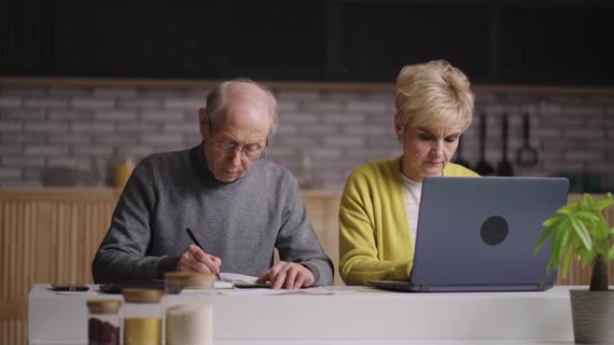 老人和女人正在支付在线水电费，一起坐在家里的厨房里，男人正在计算，女人正在使用笔记本电脑