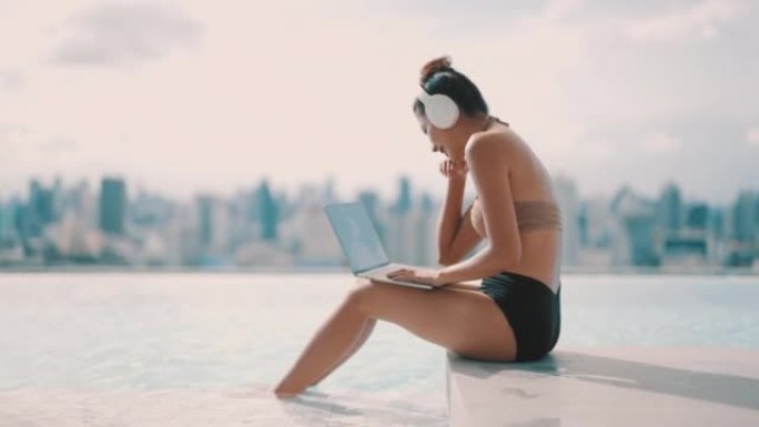 年轻女子在度假期间坐在游泳池旁，夏天在度假胜地的笔记本电脑上工作。