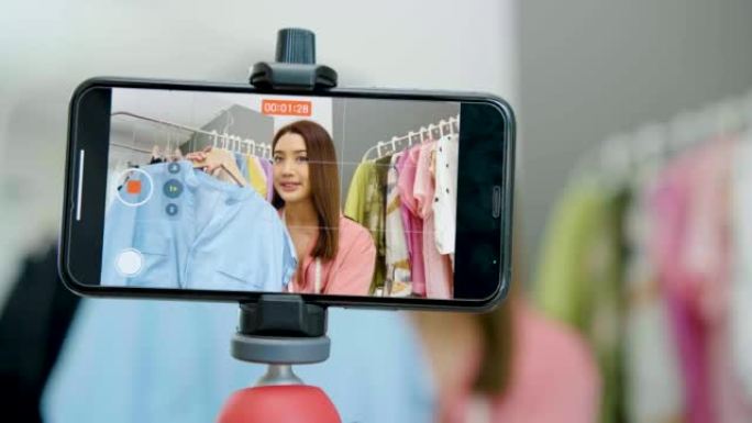 年轻的亚洲女性企业主在时装店使用智能手机直播流媒体向顾客出售时尚服装，并在社交媒体上展示细节，在线市