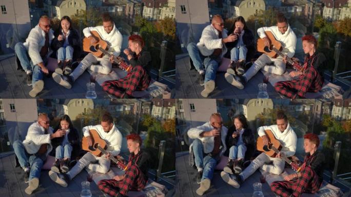宽镜头轻松的一群跨国朋友坐在屋顶上喝酒弹吉他。无忧无虑的高加索亚裔和非裔美国人在周末享受休闲。