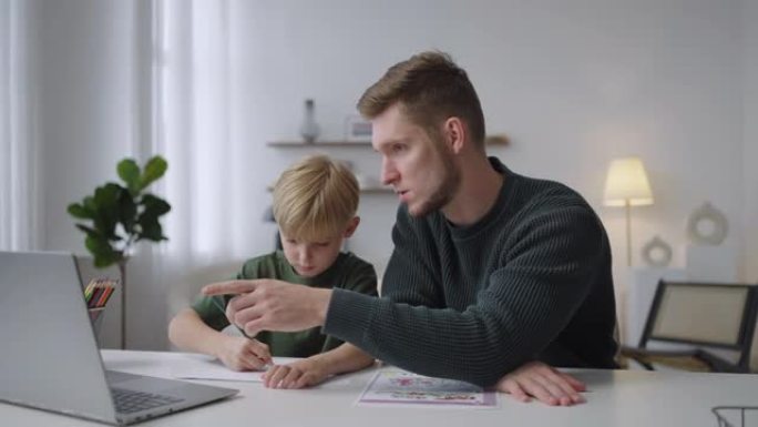 一位年轻的父亲帮助他7岁的儿子坐在客厅的桌子旁做作业。