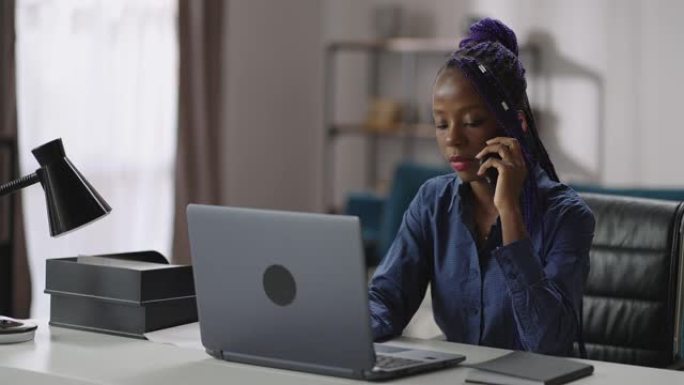 年轻的黑人女商人在家庭办公室工作，女士正在用手机聊天，坐在笔记本桌旁，咨询客户