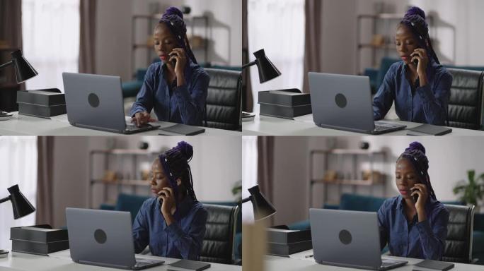 年轻的黑人女商人在家庭办公室工作，女士正在用手机聊天，坐在笔记本桌旁，咨询客户