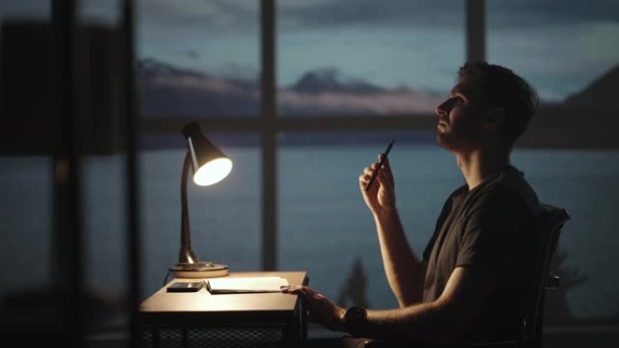 一个体贴的人坐在桌子旁，拿着台灯用笔写字。黑暗的一天黄昏在窗户的背景上一个悲伤的人思考和写作