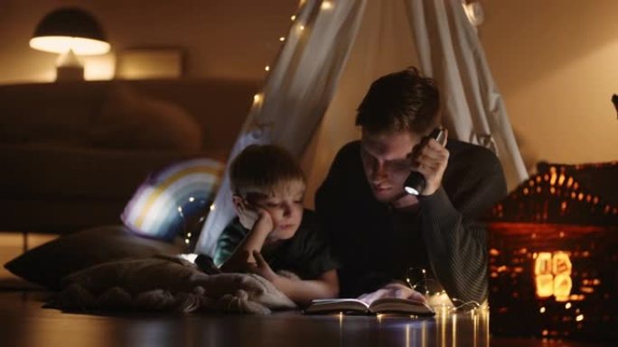细心慈爱的父亲正在家里的帐篷里为他的小儿子，爸爸和男孩读童话
