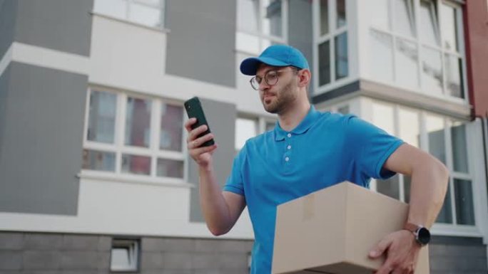 一位戴着圆形眼镜的男性邮递员，随身携带用于交付给客户的盒子，看着他的手机。通过手机搜索送货地址。支付