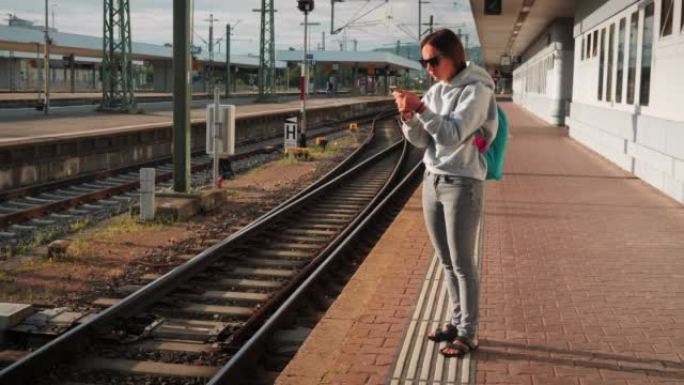 在空的铁路平台上等待火车的女人。年轻的女旅行者，背包站在空荡荡的火车站平台上，在互联网上在线寻找火车