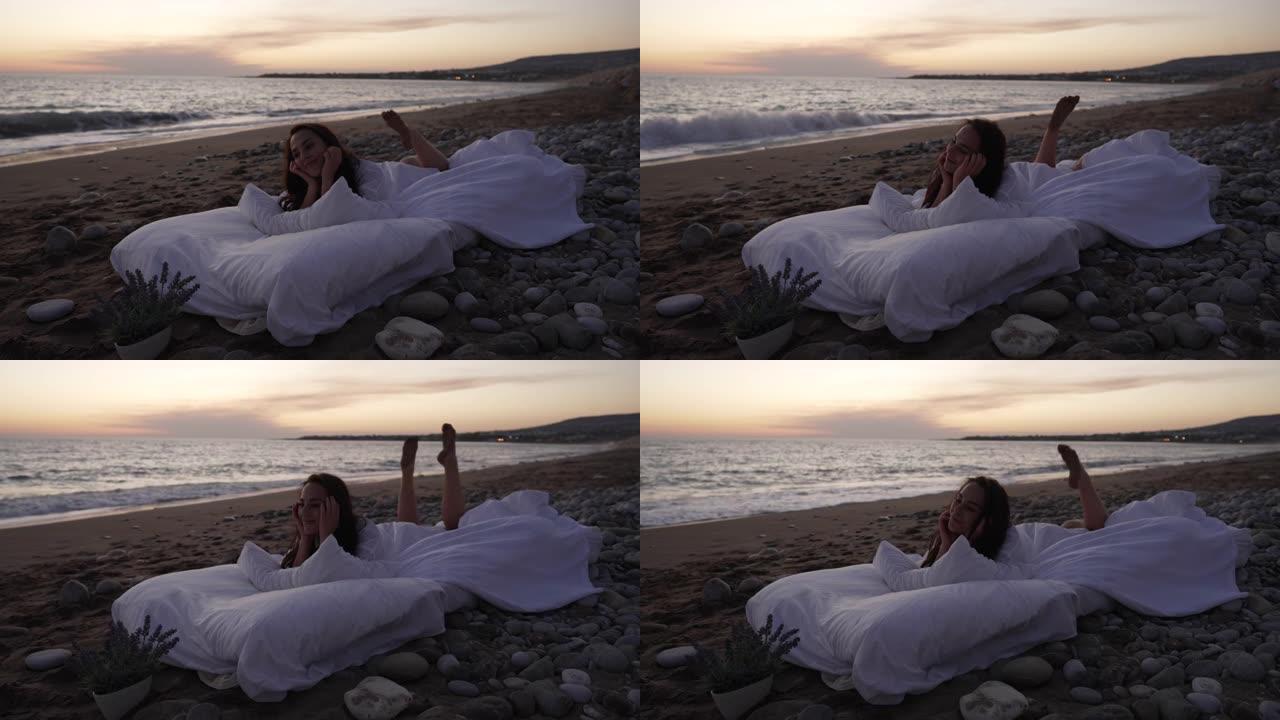 在沙滩上的暮色中，放松的年轻女子在慢动作中摇晃双腿，躺在软垫床上。快乐微笑的高加索女士在海边黄昏享受