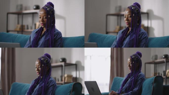 年轻的黑人妇女与紫色的辫子正在通过笔记本电脑的视频会议聊天，看着显示器并微笑着