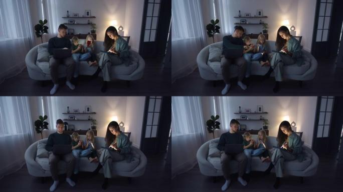 一位年轻的西班牙裔女性平面设计师与家人坐在沙发上时正在平板电脑上工作。孩子们玩手机。丈夫在笔记本电脑