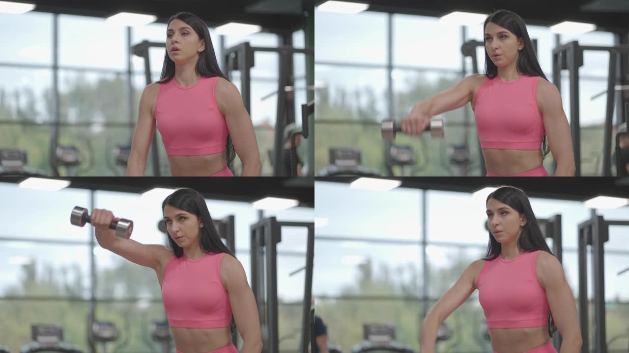 穿着粉红色西装的黑发女人在健身房训练肩膀时，交替将哑铃抬高到她面前的顶部。训练肩膀和手臂的站立练习。
