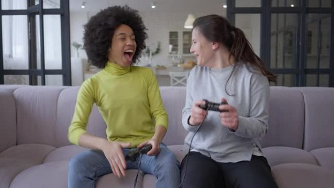年轻快乐的女性游戏玩家给高五玩多人视频游戏的肖像。快乐无忧无虑的高加索人和非洲裔美国朋友坐在室内的沙