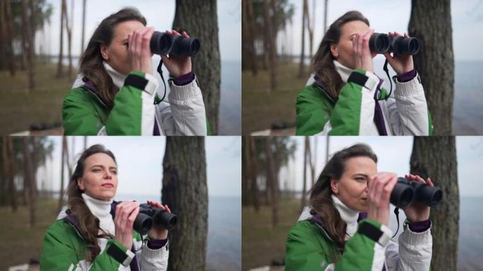 自信的女性旅行者站在林地的河岸上，用双筒望远镜将目光移开。春秋日户外旅行的高加索美女肖像享受徒步旅行