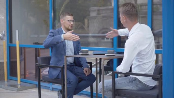 两个快乐的高加索商人在户外咖啡馆的餐桌旁握手交谈。成功自信的男人在午餐时做生意。商业和成功。