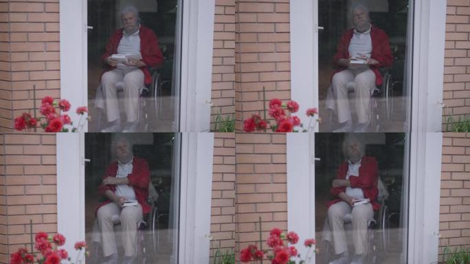 坐在轮椅上的自信聪明的英俊老头看书，思考坐在家里的玻璃门后面。聪明体贴的高加索退休人员在室内的宽幅肖