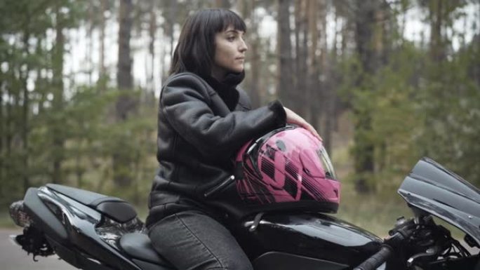 自信微笑的白人妇女坐在黑色摩托车上，戴着粉红色头盔，看着远处的侧视图。美丽的年轻女摩托车手在户外思考