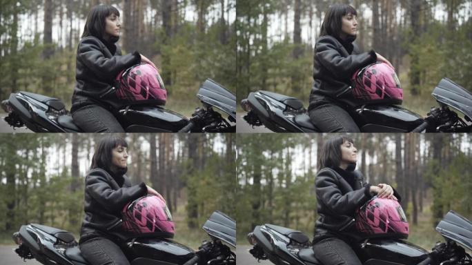 自信微笑的白人妇女坐在黑色摩托车上，戴着粉红色头盔，看着远处的侧视图。美丽的年轻女摩托车手在户外思考