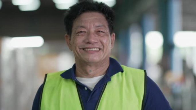 职业亚裔男工身穿安全服，手持安全帽站在仓库里，笑嘻嘻地看着镜头的画像。在产品配送物流中心或制造仓储工